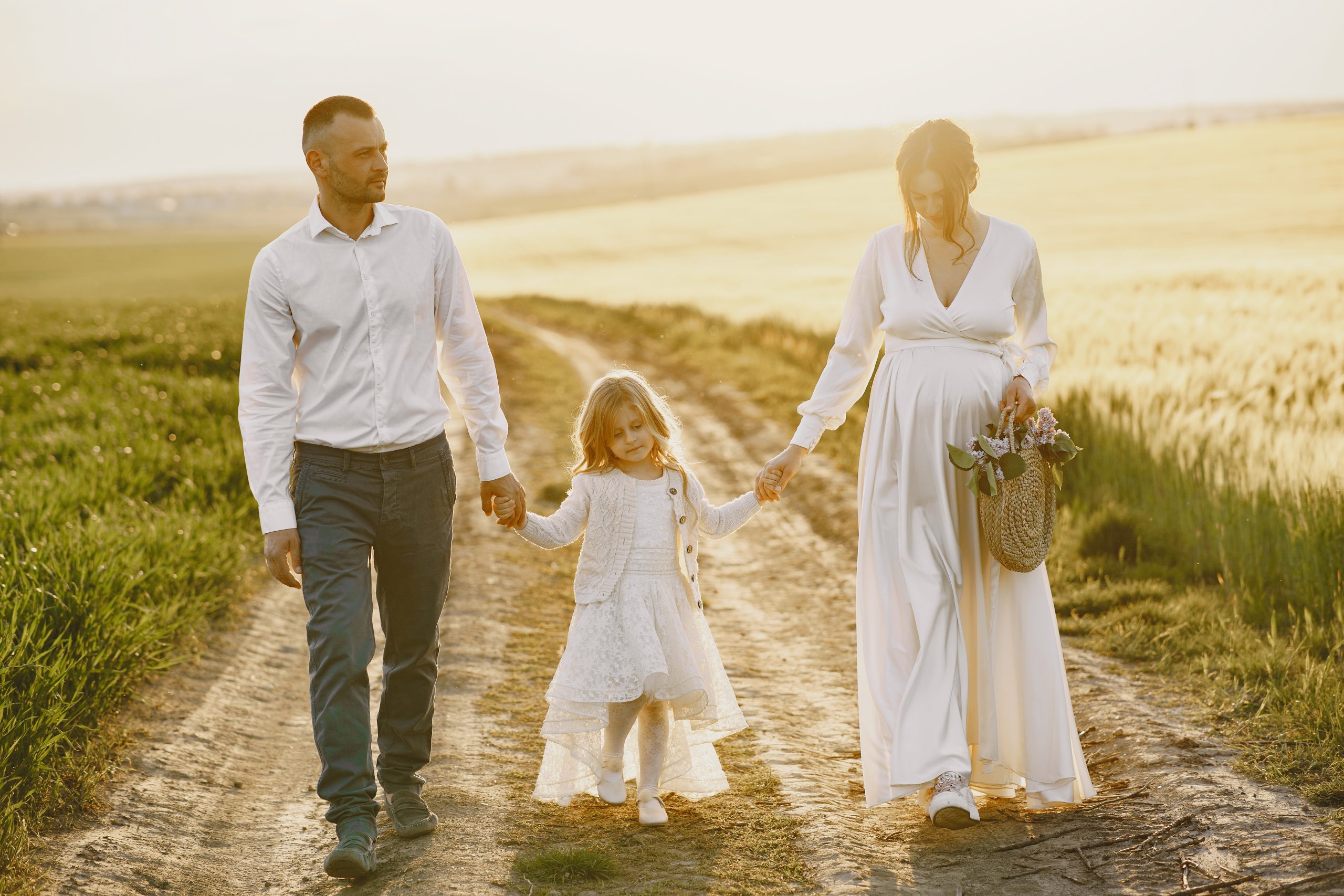 Pregnant mother and daughter. Little girl child in the summer meadow. Woman in a white dress. Sunset background.